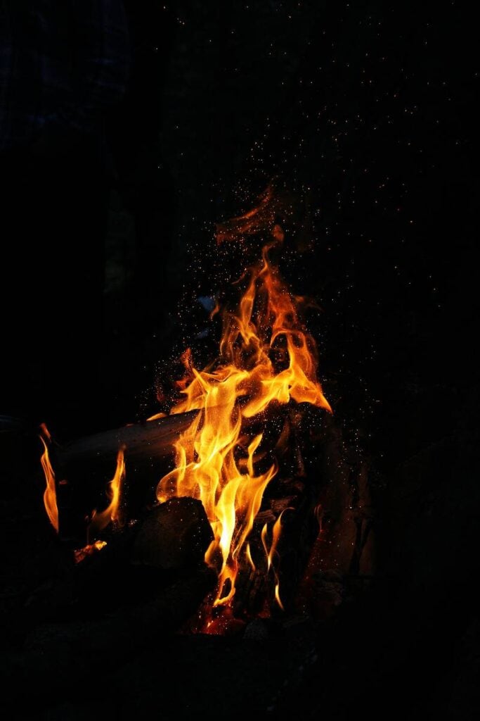 Close-up of a glowing campfire with vivid orange and yellow flames rising from stacked logs against a dark background. Tiny sparks float in the air, creating a warm and atmospheric scene.