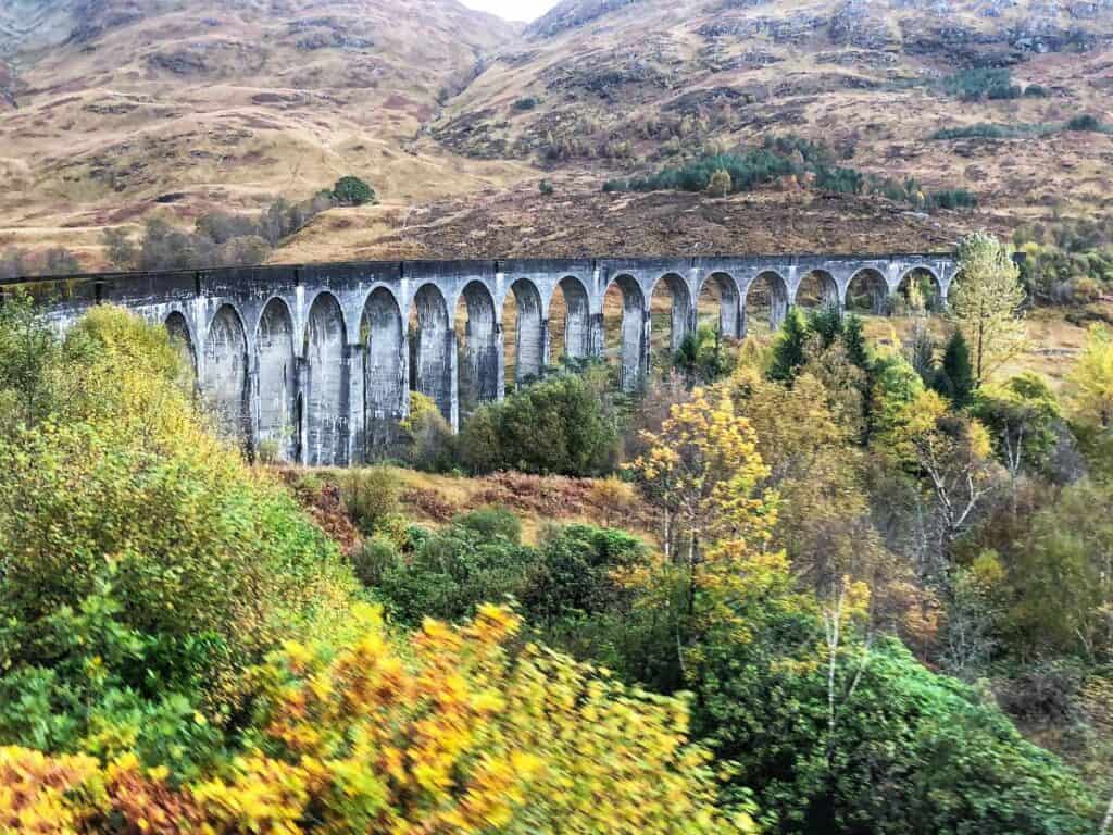The Glenfinnan Viaduct, a sweeping stone railway bridge with 21 tall arches, stretches across a lush valley surrounded by autumn-colored trees and rolling Scottish hills. The iconic structure curves gracefully through the landscape, with weathered gray stone contrasting against the golden and green foliage.