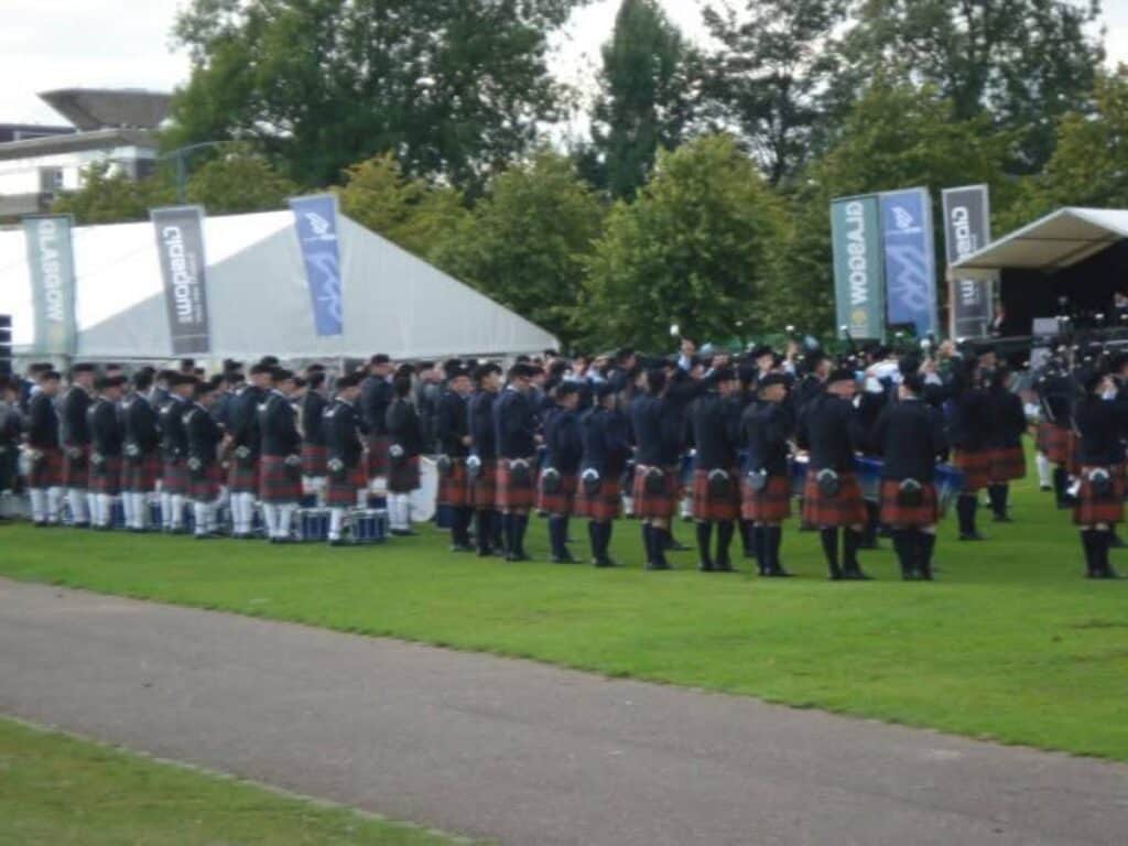 Large group of people dressed in traditional Scottish attire, including kilts and sporrans, gathered on a grassy field during a cultural or ceremonial event. Tents, banners labeled "Glasgow" and "Worlds," and a stage with musicians suggest this is a competitive pipe band gathering or festival in Scotland.