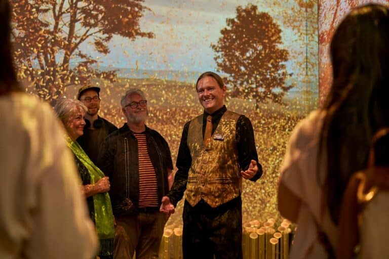 A smiling man in a gold vest and tie speaks to a small group of people in a warmly lit room with a vivid autumn-themed backdrop showing trees and falling leaves. The group listens attentively, some smiling, as golden light and shadows from the projection create a cozy, immersive atmosphere.