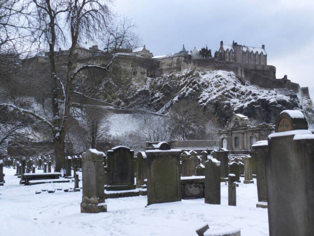 Snow blankets the historic gravestones in St Cuthbert's Churchyard beneath Edinburgh Castle on an overcast winter day. The castle rises dramatically above the cemetery while weathered headstones and monuments create a peaceful, atmospheric scene in the heart of Edinburgh.